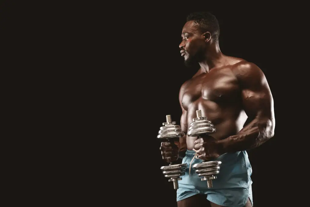 A photograph of a muscular athletic black male with dumbells in his hands. Strong lighting from the sides highlights his physicque.