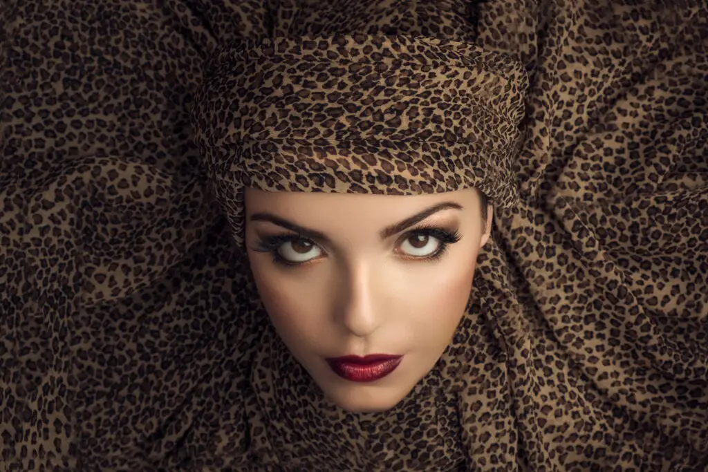 Beauty shot of a woman with finely textured skin, deep red lips and large eyelashes looking upwards towards the camera. The background is a headband made of leopard skin pattern cloth which frames her entire face.