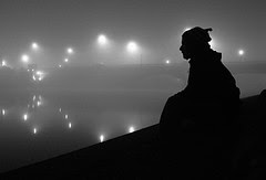 A night-time portrait of a man silhouetted against fog and lights in the background, with a water reflection. Low Light Photography is a challenge. 